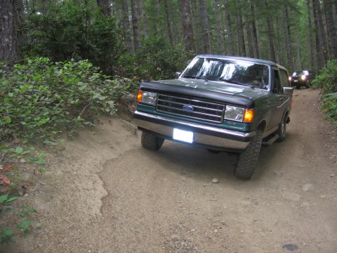 1990 Ford Bronco - A green 1990 Ford Bronco navigates a wooded dirt road, with another vehicle visible in the distance.