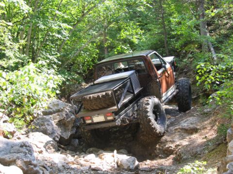1981 Ford Bronco - Pickup truck with custom rear rack and spare tire drives off-road on a steep, rocky path.