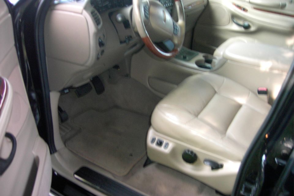 Beige leather car interior showing driver seat, wood-trimmed steering wheel, pedals and floor mat