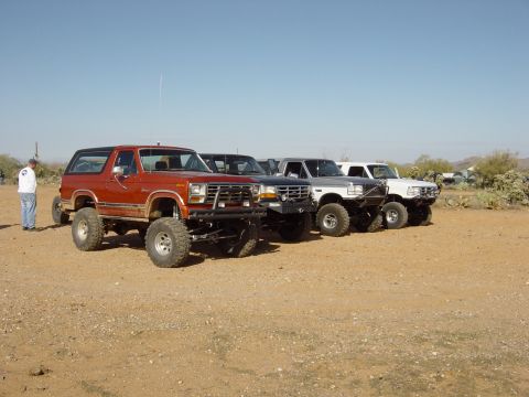 1993 Ford Bronco - A line of lifted SUVs, including a red one with a custom bumper, are parked in a desert setting under a clear blue sky.