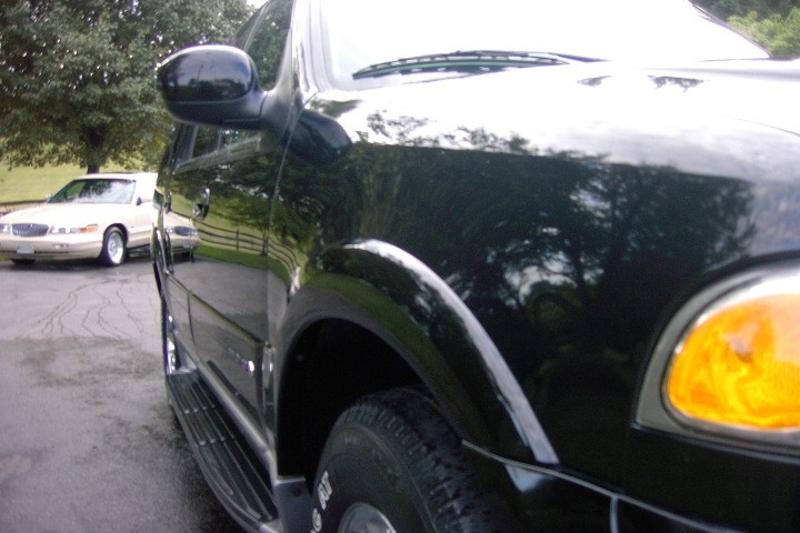 Close-up along a dark green SUV's front fender with a white full-size sedan parked in the background