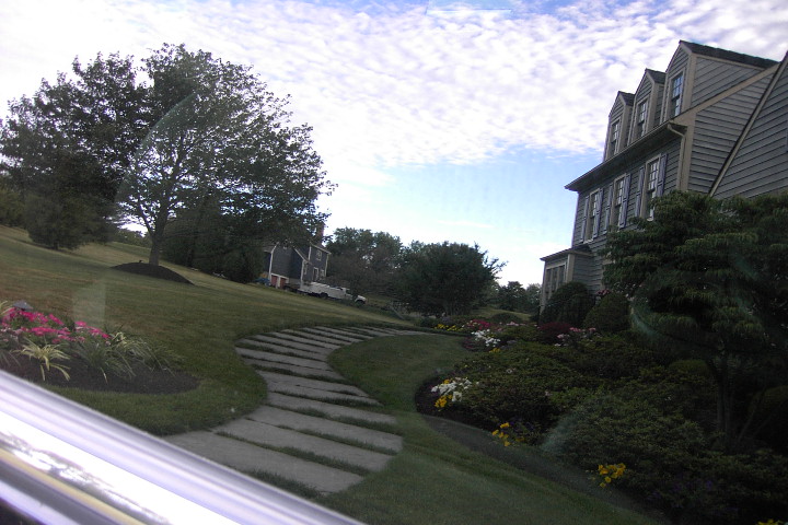 Landscaped lawn with stone path and flower beds beside a large gray house, seen through a car window