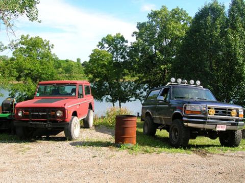 1994 Ford Bronco - Two Ford Broncos are parked near a body of water under a partly cloudy sky.