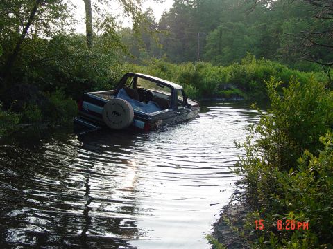 1995 Dodge 1500 - A dark blue pickup truck is stuck in a stream, with its spare tire visible and the water reaching the truck bed.