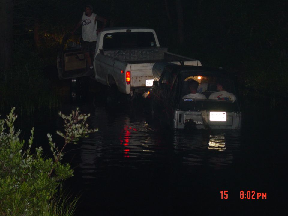Two trucks are partially submerged in water at night. One truck is white, the other is dark. People are visible inside the dark truck