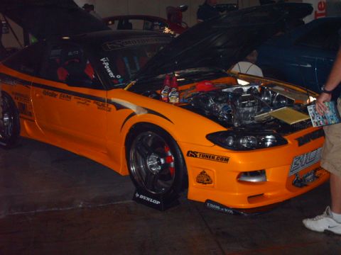 1995 Ford Probe - An orange sports car is displayed with its hood open, revealing the engine and numerous sponsor decals on its body.