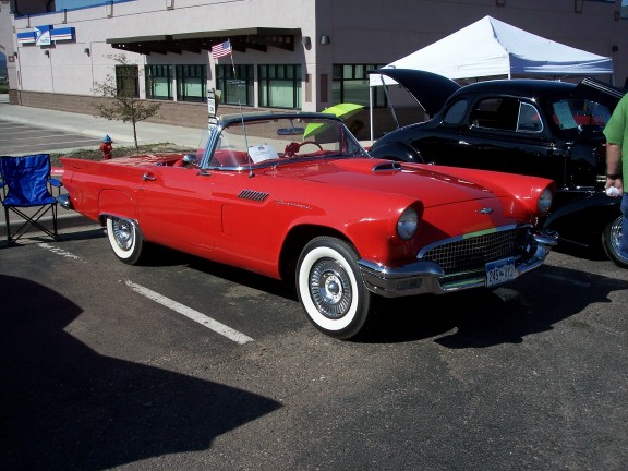 Red convertible with white-wall tires and chrome grille. A black car is parked behind it