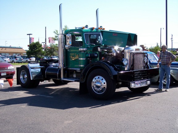 Green semi-truck with chrome stacks and grille, hood open, parked at an event