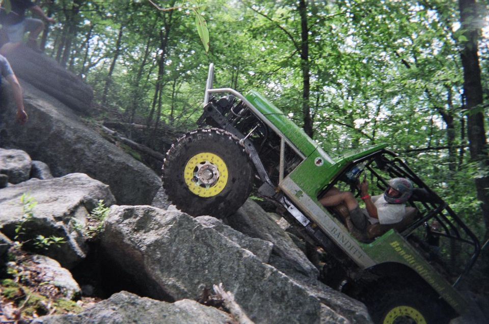 Green off-road buggy with roll cage climbing over large rocks on a wooded trail. Driver wears a helmet