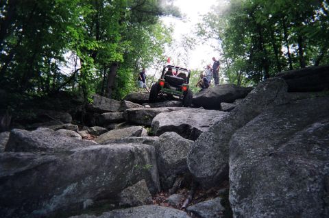 1976 GMC C/K Pickup - An off-road vehicle tackles a challenging rocky trail surrounded by trees.