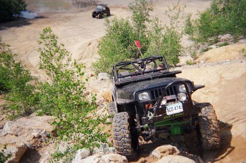 1976 GMC C/K Pickup - An off-road vehicle navigates a dusty trail surrounded by greenery, with another vehicle in the distance.