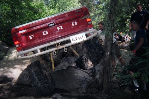 1976 GMC C/K Pickup - A red Ford pickup truck is shown off-roading through a rocky, wooded area, with several people observing.