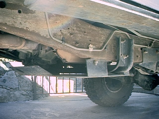 Undercarriage of a pickup showing frame, fuel tank brackets, lines, and a rear tire with off-road tread