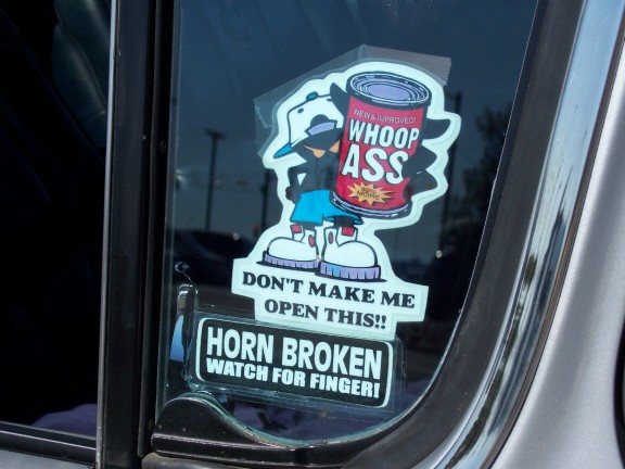 Car window with 'Whoop Ass' sticker and 'Horn Broken Watch For Finger' sticker