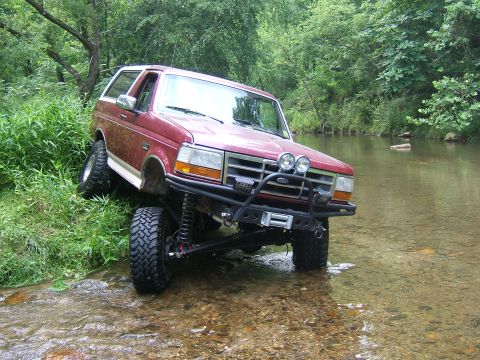 1993 Ford Bronco - A red SUV navigates through a watery path surrounded by lush greenery.