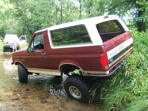 1993 Ford Bronco - A maroon and white Ford Bronco navigates a stream, with another vehicle visible in the background.