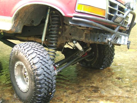 1993 Ford Bronco - Close-up of the front suspension and off-road tire of a red 1993 Ford Bronco, partially submerged in shallow water.