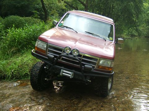 1993 Ford Bronco - This red SUV is navigating a rocky creek bed surrounded by lush green foliage.