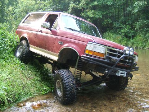1993 Ford Bronco - Red 1993 Ford Bronco navigating a rocky stream bed with its tires partially submerged.
