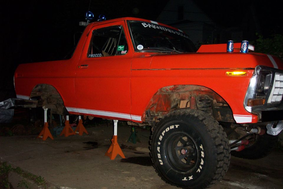 Orange truck on jack stands, hood open, two cans on hood. Windshield banner says 'BARN FIND BUILD'. Door says 'PROCCO'. Tires say 'USA SPORT TRAIL-X'