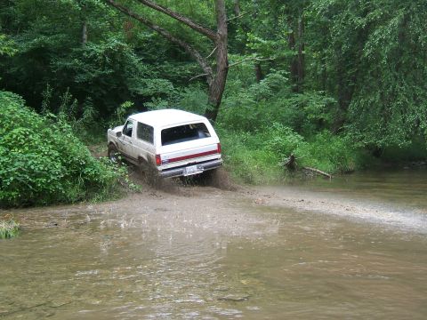 1993 Ford Bronco - A white SUV navigates a challenging off-road path, kicking up mud and water.
