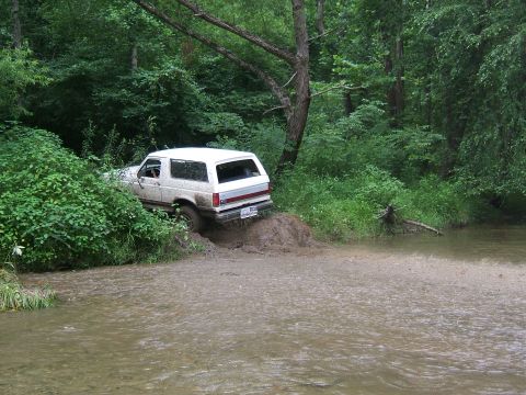 1993 Ford Bronco - White SUV navigates a muddy creek bank, its rear wheels sinking into the mire.