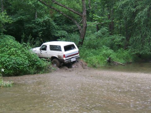 1993 Ford Bronco - A white 1993 Ford Bronco navigates a muddy creek crossing, with trees and foliage surrounding the scene.