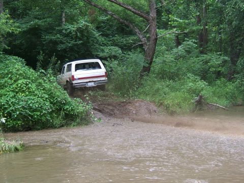 1993 Ford Bronco - A white SUV navigates a muddy trail and stream in a wooded area.