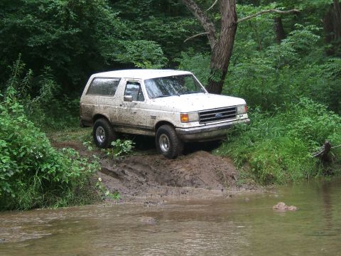 1993 Ford Bronco - A white Ford Bronco navigates a muddy trail next to a shallow stream in a wooded area.