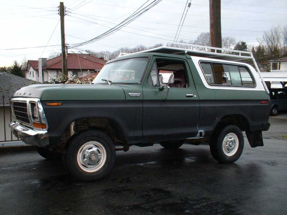 Green 1979 Ford Bronco with white top and roof rack, driver-side front 3/4 view on wet pavement