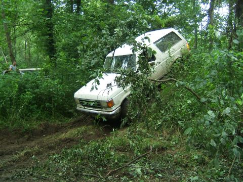1993 Ford Bronco - A white SUV is stuck in the mud and foliage in a wooded area, with another vehicle partially visible in the background.