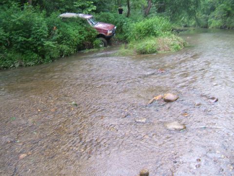 1993 Ford Bronco - A red SUV is parked off-road next to a stream, surrounded by dense green foliage.
