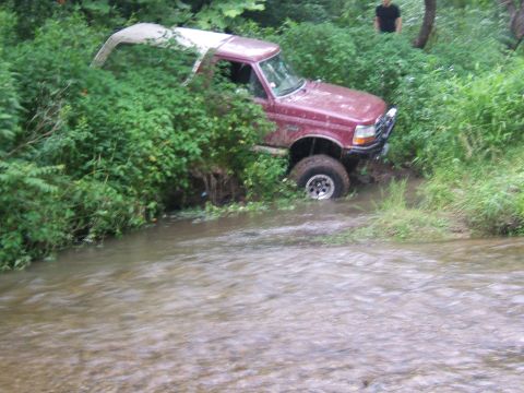 1993 Ford Bronco - A red SUV is stuck in a creek, partially submerged in water and surrounded by dense foliage.