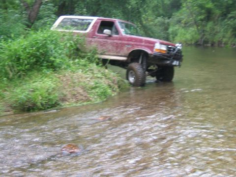 1993 Ford Bronco - A red SUV with a white top navigates a shallow creek, its lifted suspension and off-road tires visible.
