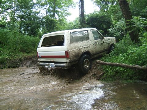 1993 Ford Bronco - Ford Bronco driving off-road through a muddy creek bed surrounded by dense green foliage.