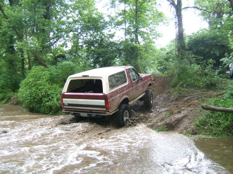 1993 Ford Bronco - Maroon and white Ford Bronco navigates a muddy stream with a lifted suspension.