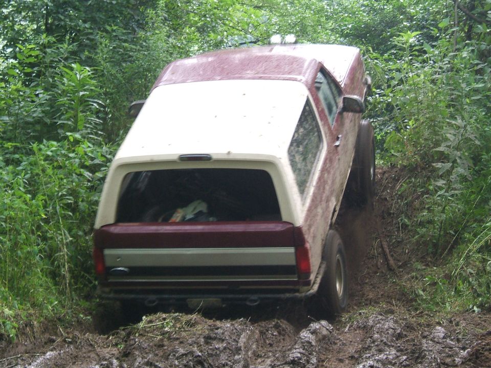 Rear view of a maroon and white Ford Bronco driving through a muddy trail with trees on both sides
