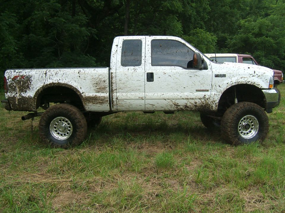 Mud-splattered white pickup truck with lifted suspension and large tires, parked on grass