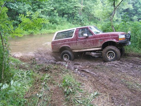 1993 Ford Bronco - A maroon 1993 Ford Bronco navigates a muddy off-road trail with water crossing.