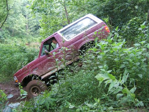 1993 Ford Bronco - A red 1993 Ford Bronco is shown navigating a steep, muddy off-road trail surrounded by dense foliage.