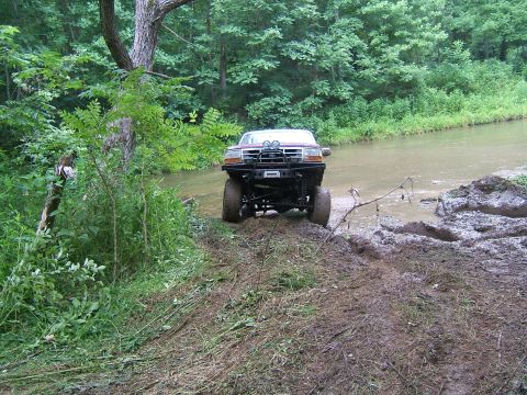 1993 Ford Bronco - A lifted Ford truck navigates a muddy creek bed surrounded by dense forest.
