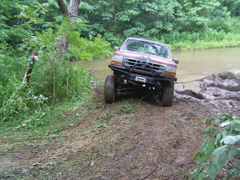 1993 Ford Bronco - A red Ford Bronco navigates a muddy bank next to a body of water, its front winch engaged.