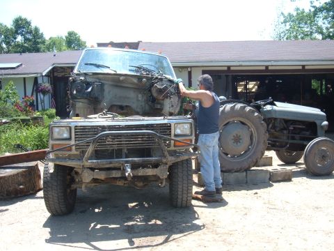1985 Ford F-150 - A man works on a truck chassis that has had its engine removed, with a tractor visible behind it.
