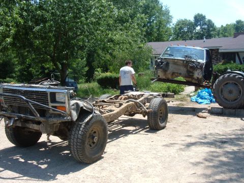 1985 Ford F-150 - A truck chassis sits in the foreground while its cab is lifted by a tractor in the background.