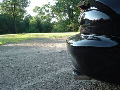 2002 Chevrolet Camaro - The rear of a black 2002 Chevrolet Camaro is visible, parked outdoors on a gravel road with trees in the background.