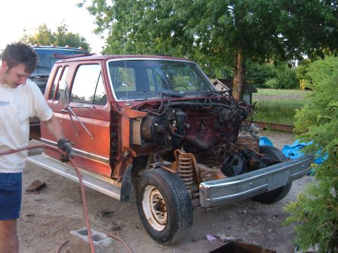 1985 Ford F-150 - A person works on a red extended cab truck that has had its front end disassembled, revealing the engine bay and suspension components.