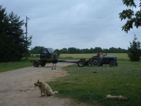 1985 Ford F-150 - A tractor is used to tow a truck chassis across a rural landscape under a cloudy sky.