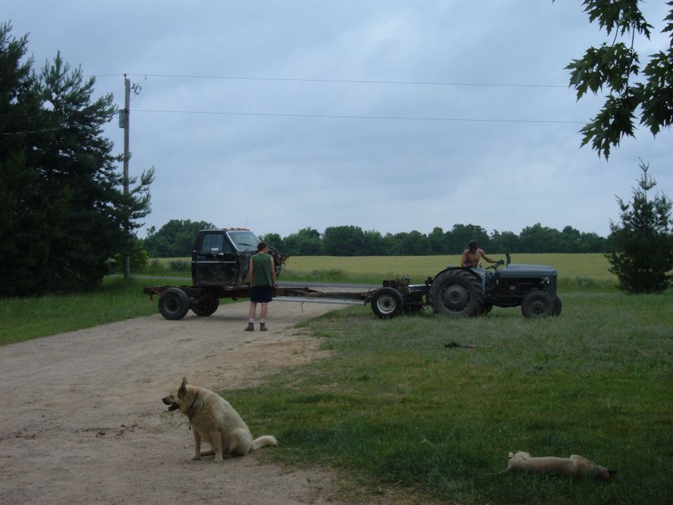 Tractor pulling a truck chassis down a dirt road, with a dog sitting nearby
