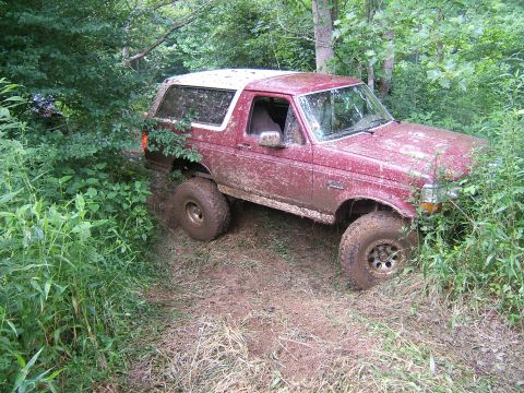 1993 Ford Bronco - This red SUV is driving through a wooded area, covered in mud from the trail.