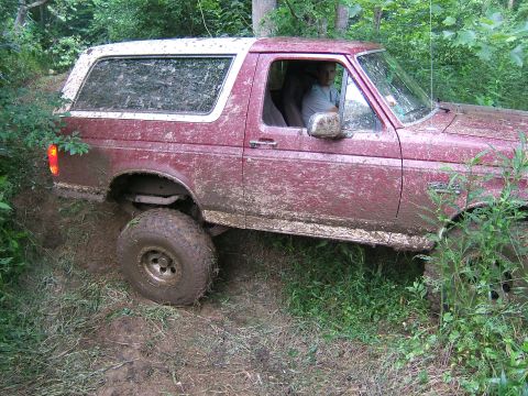 1993 Ford Bronco - Mud-covered maroon 1992 Ford Bronco with a white top, driving off-road through a wooded area.
